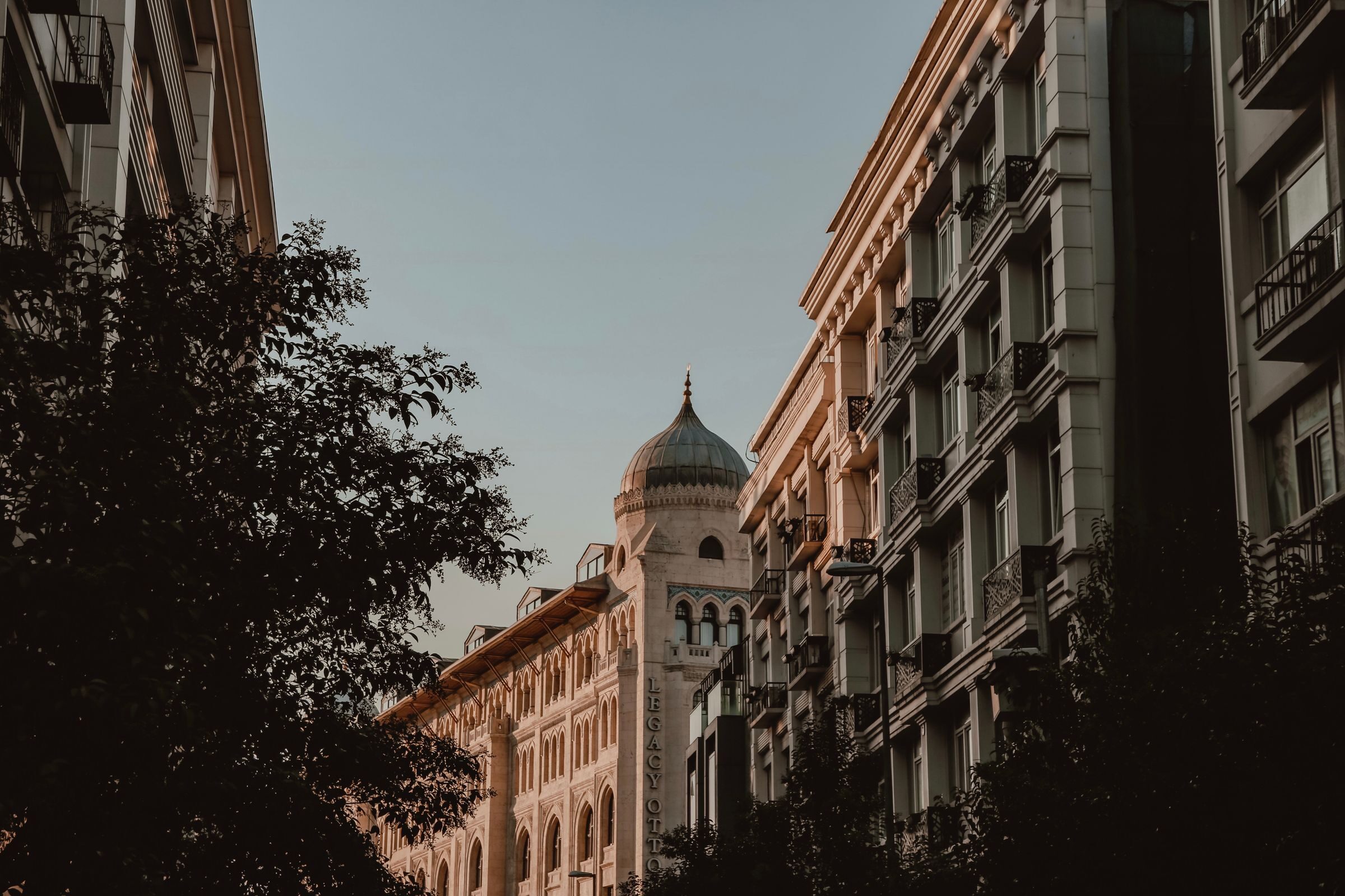 Sunlit stone and cream-coloured apartment buildings along a tree-lined street, with a domed tower rising in the distance against a pale evening sky.