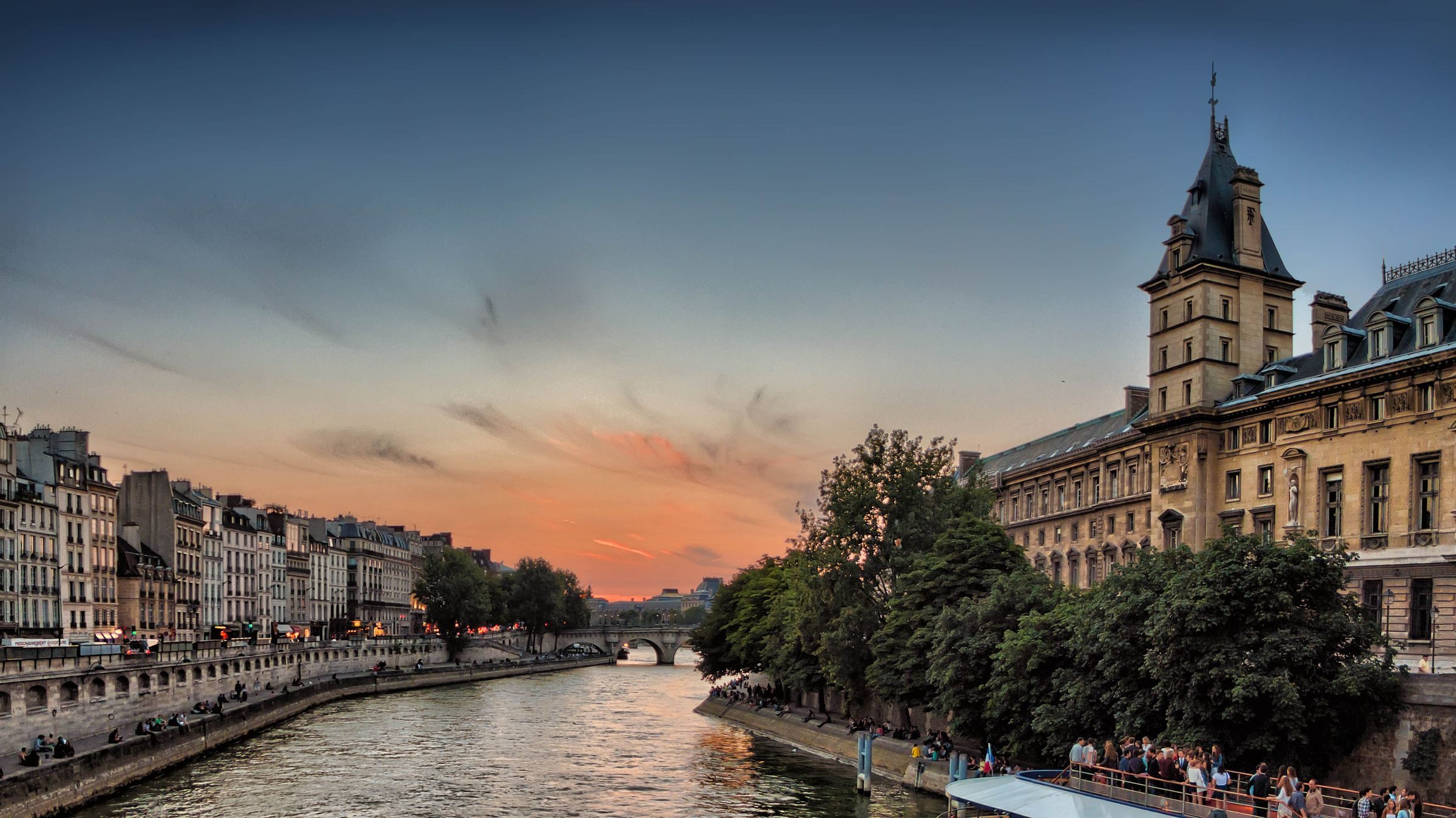 A river at sunset between stone embankments, with historic buildings, trees and a bridge in the distance.